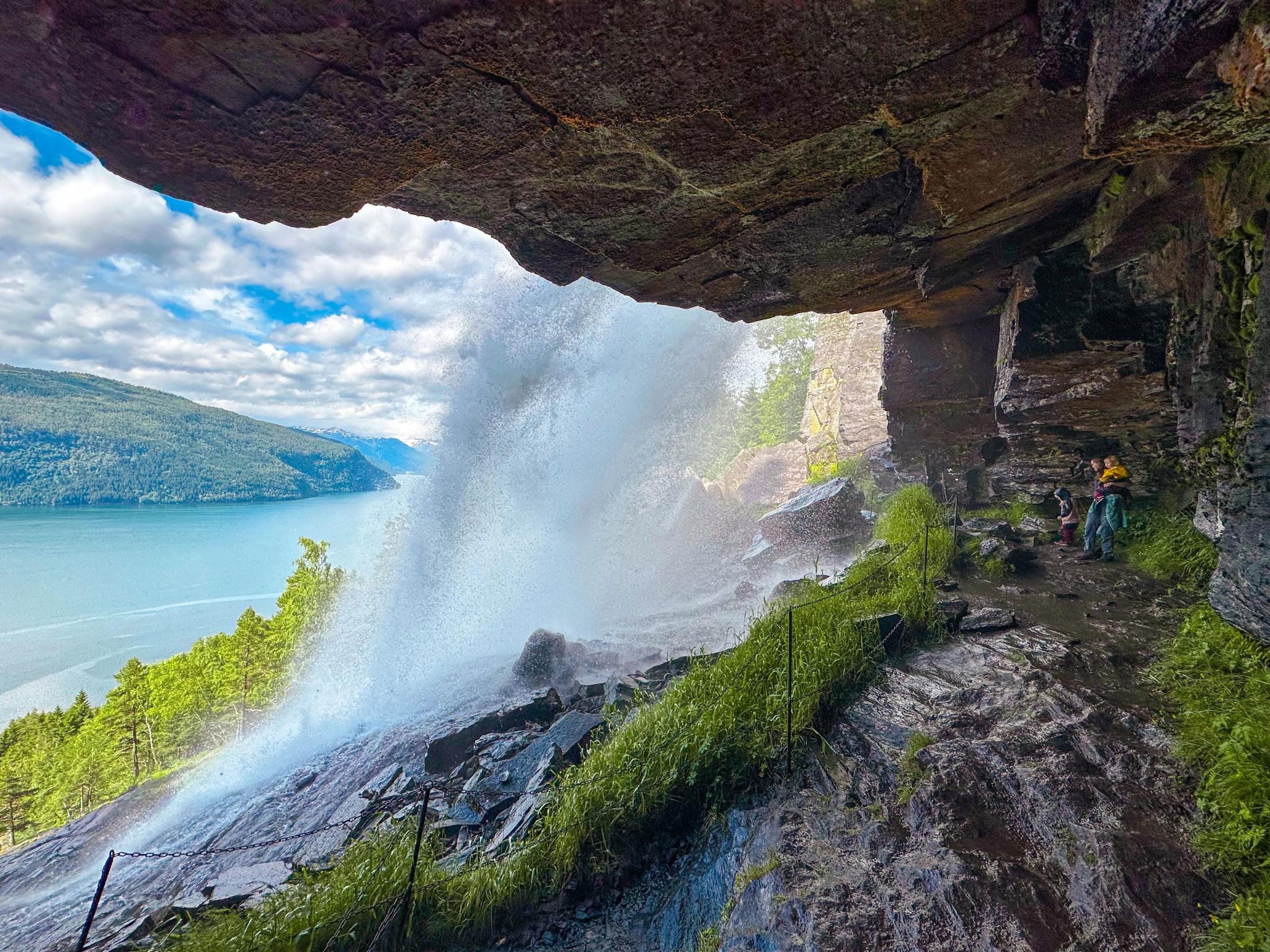 View from under a rocky overhang beside a misty waterfall, with hikers on a wet path and a lake beyond.