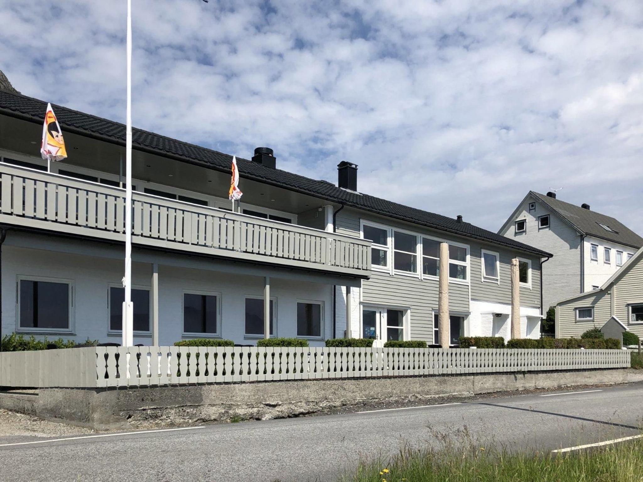 Two-story gray building with balcony, flags and fenced yard beside a paved road under a partly cloudy sky..