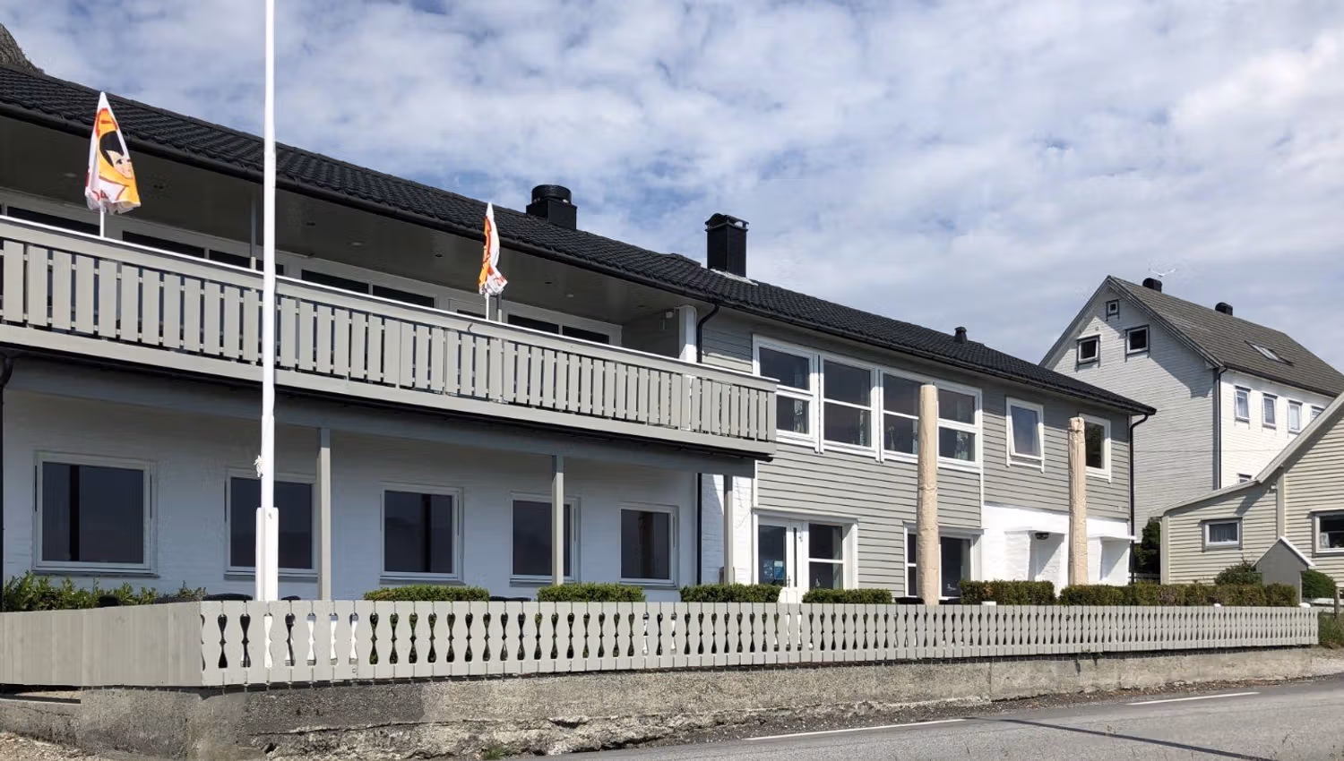 Two-story gray building with balcony, flags and fenced yard beside a paved road under a partly cloudy sky..