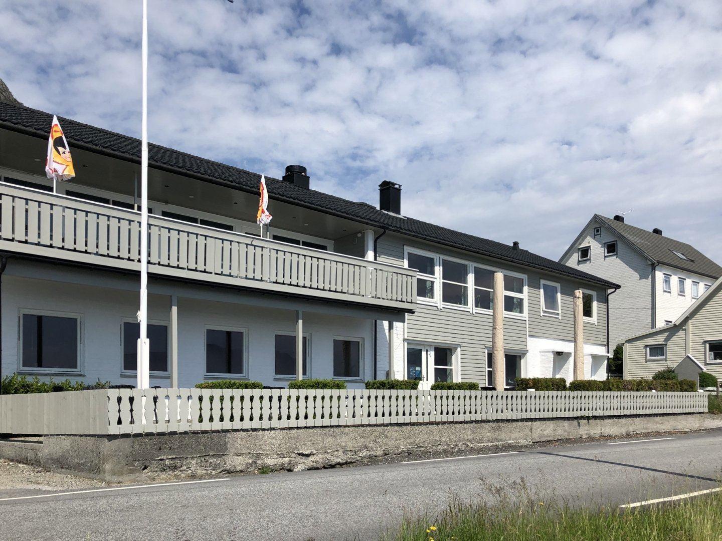 Two-story gray building with balcony, flags and fenced yard beside a paved road under a partly cloudy sky..