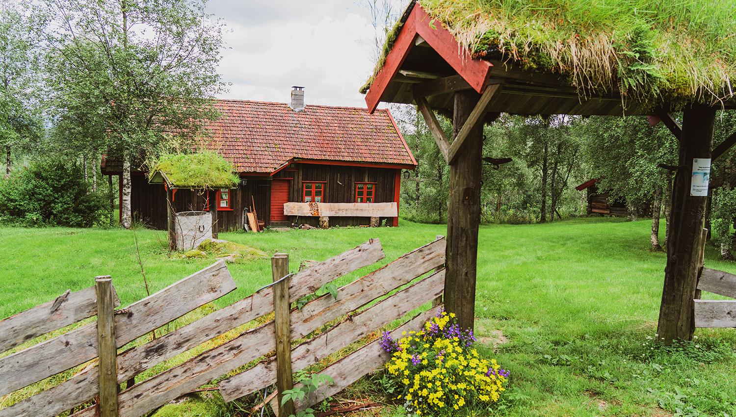 A wooden garden arch with a turf roof, a large grassy area with a brown cabin featuring red trim on the moldings and door.