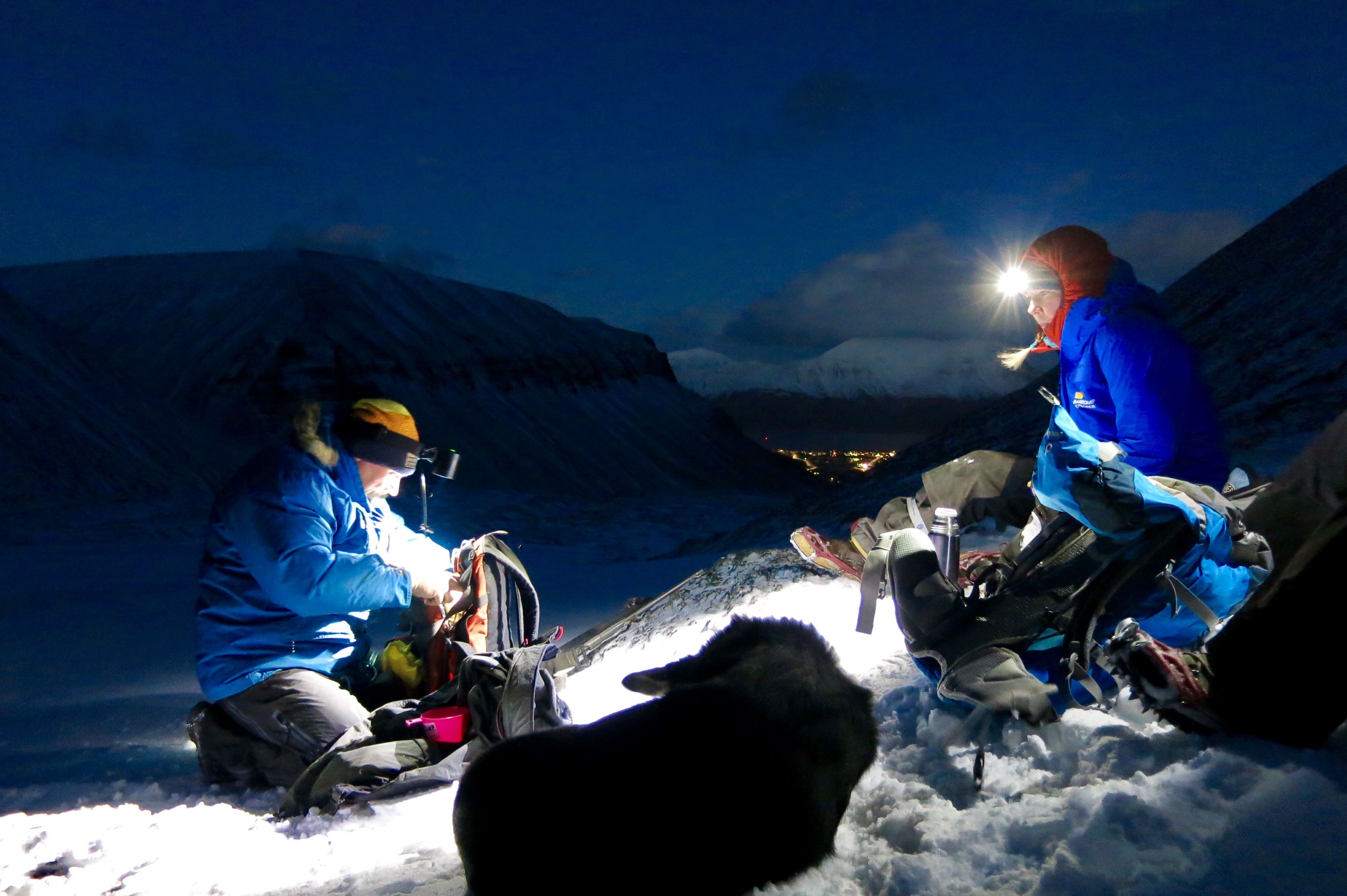 To personer med hodelykt og en hund i forgrunnen, med fjell og Longyearbyen i bakgrunnen.