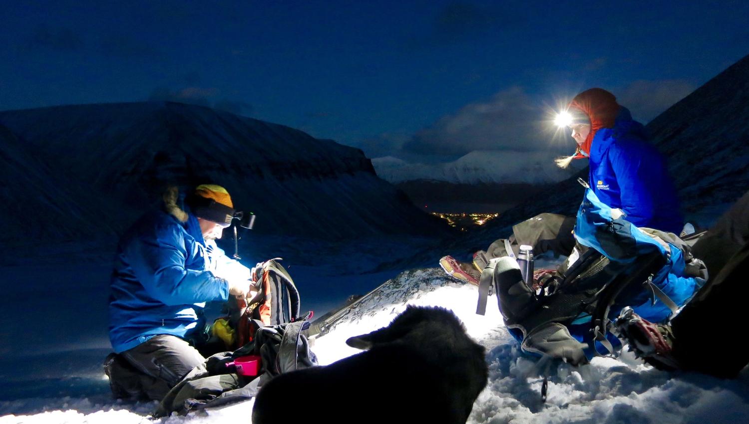 To personer med hodelykt og en hund i forgrunnen, med fjell og Longyearbyen i bakgrunnen.