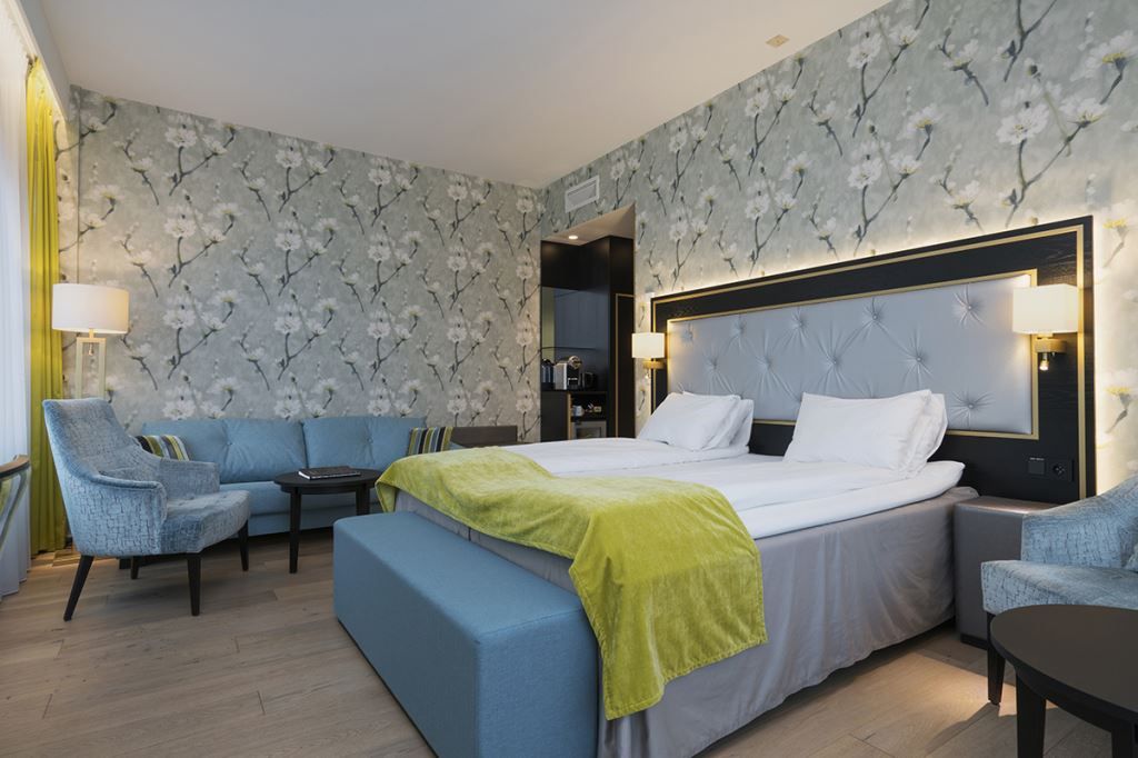 Interiør av hotellrom: Dobbeltseng, blå lenestol og sofa, blomstret tapet.