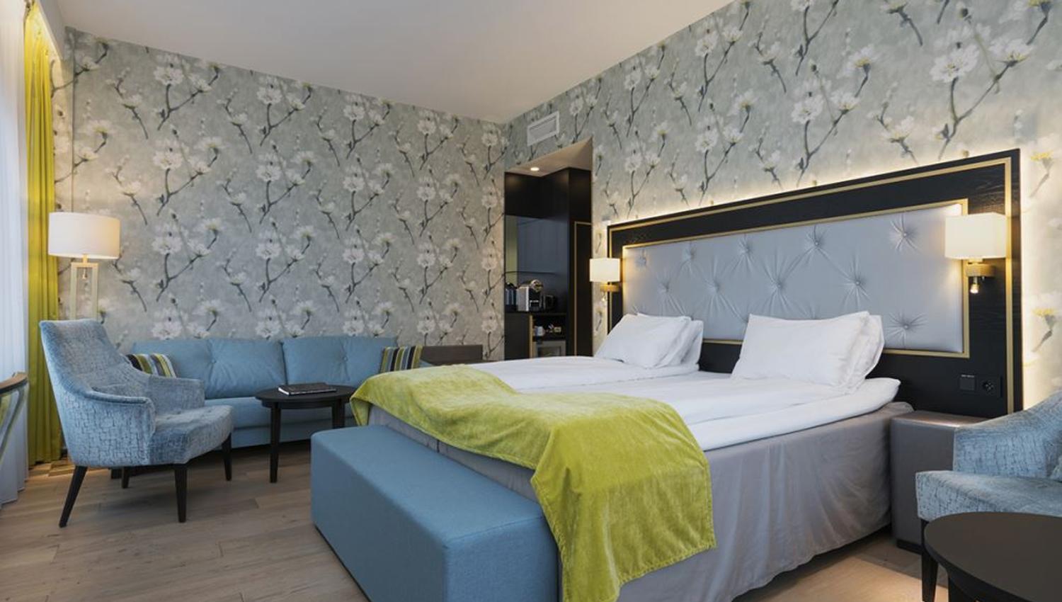 Interiør av hotellrom: Dobbeltseng, blå lenestol og sofa, blomstret tapet.