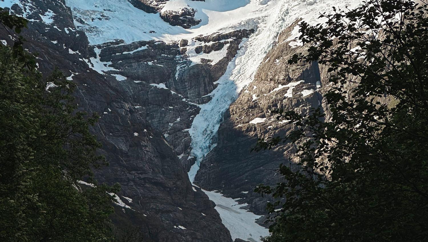 Snow and ice cling to a rugged mountain face above a dark green forested valley under a clear blue sky..