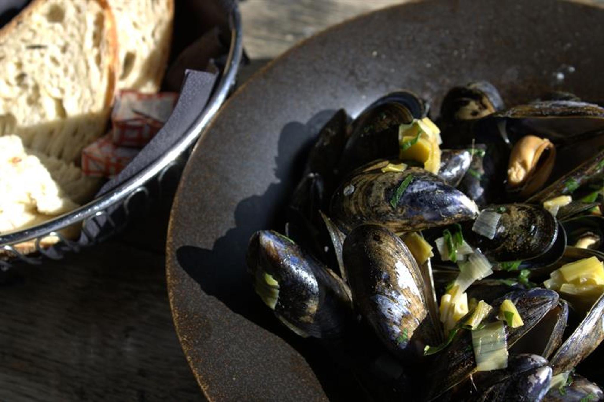 Mussels served in a deep bowl with herbs and sauce, accompanied by bread. The dish is suitable for sharing or as a main course.