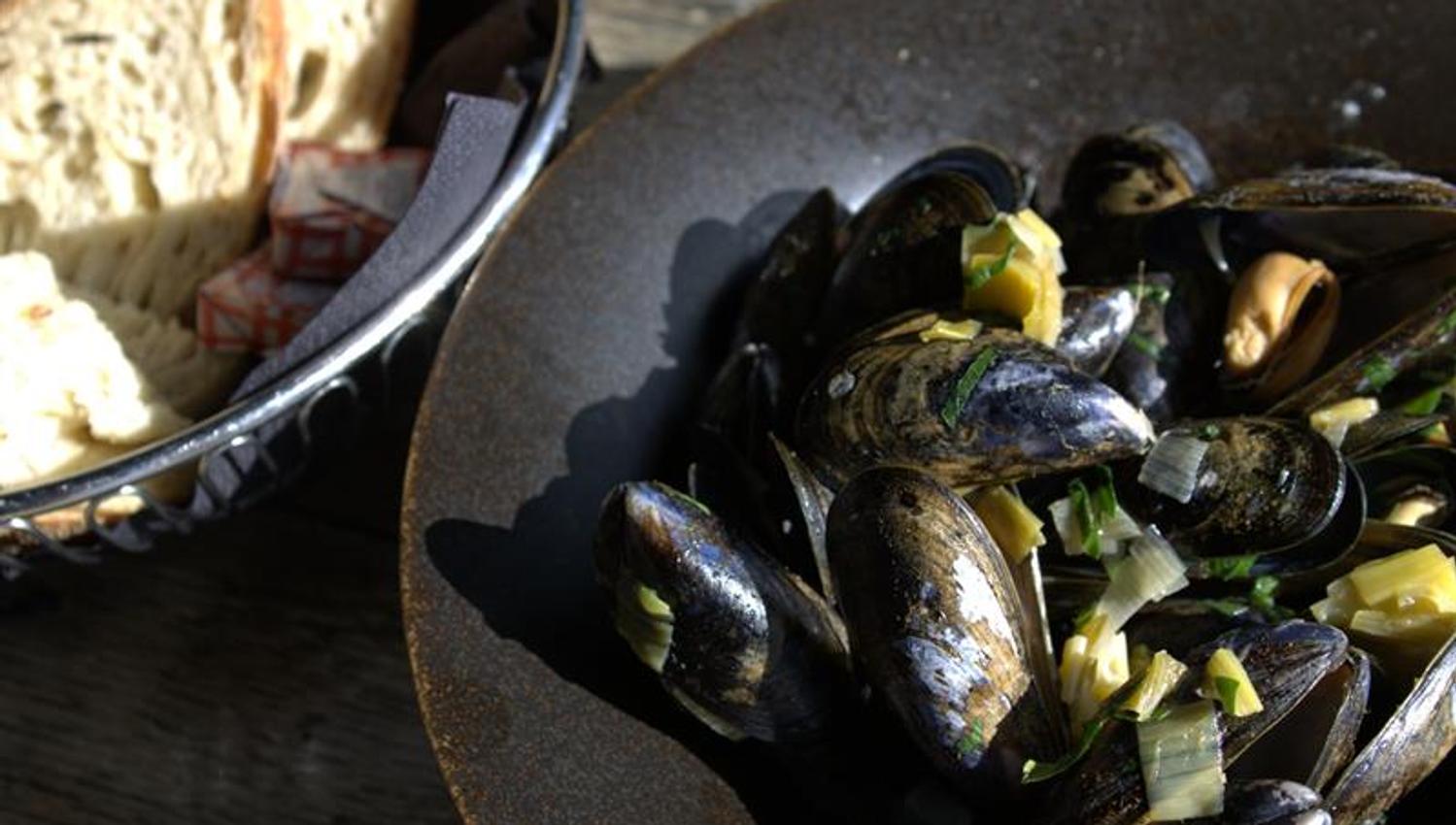 Mussels served in a deep bowl with herbs and sauce, accompanied by bread. The dish is suitable for sharing or as a main course.