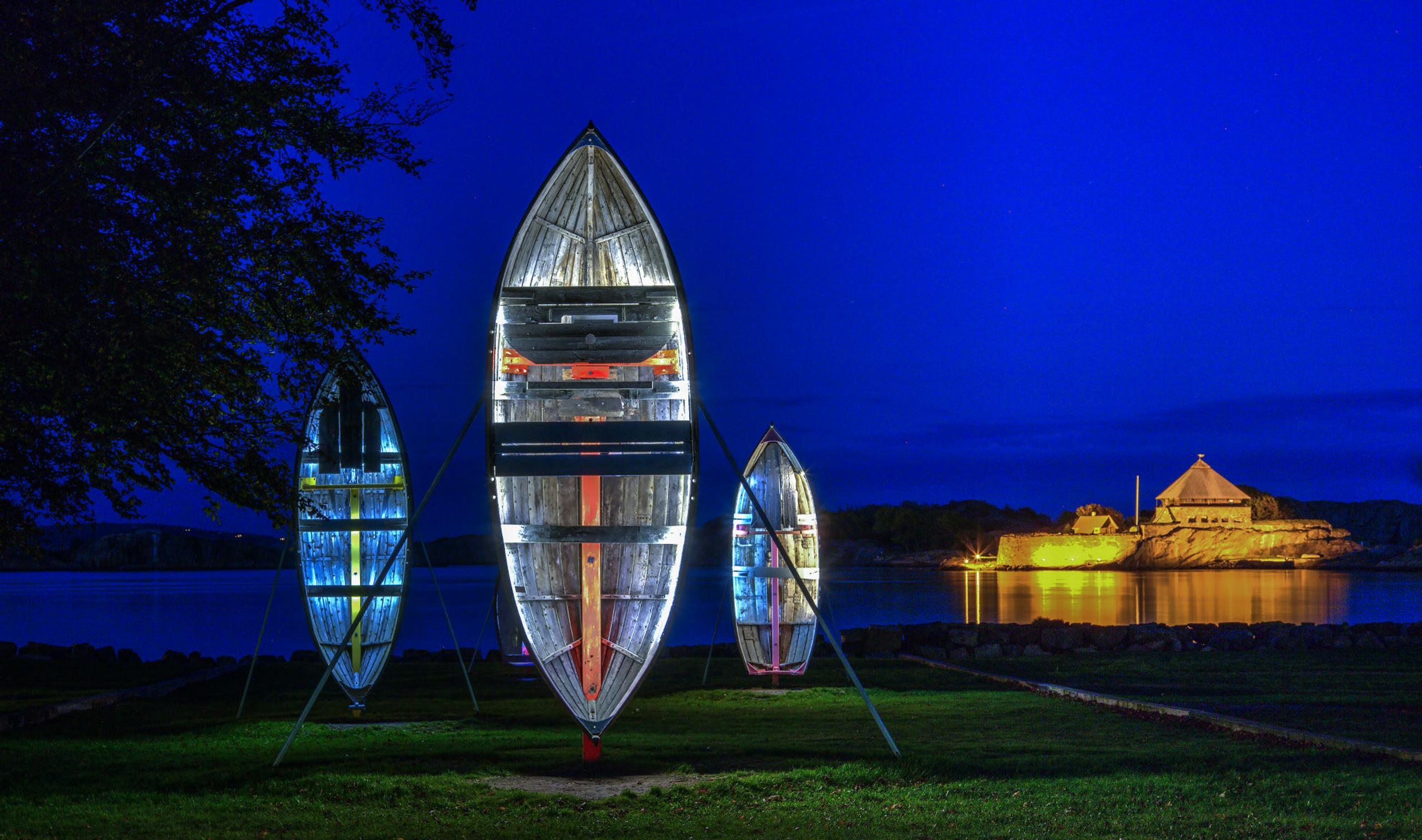 Three illuminated boat-shaped art installations by the sea, with the Citadel in the background.