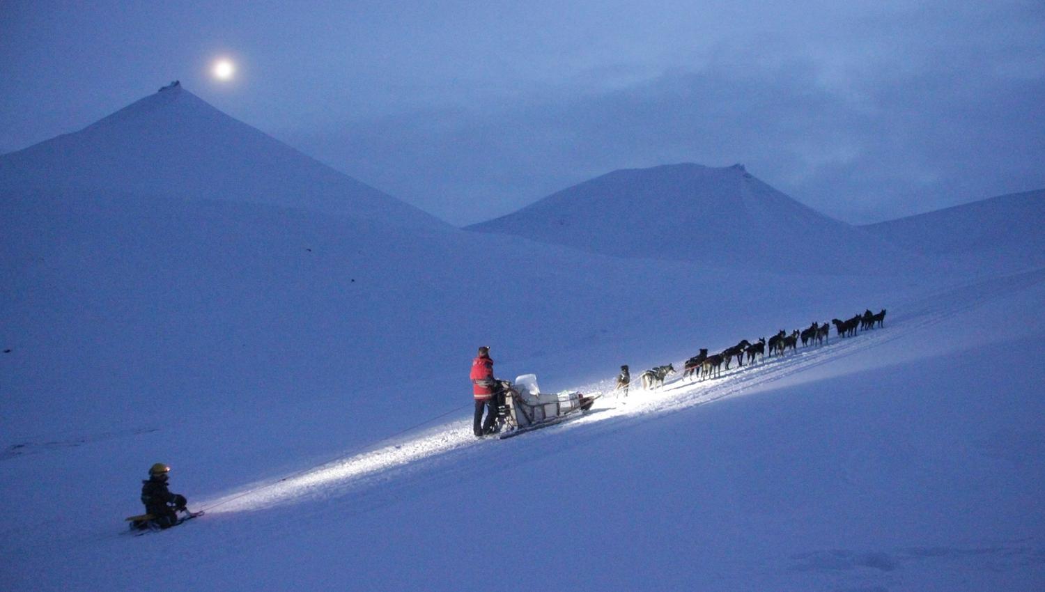 A person driving a dog sled with a child being towed on a children's sled behind