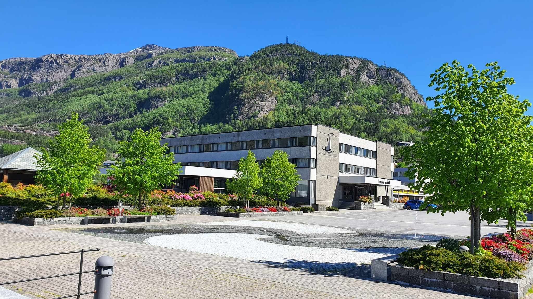 Moderne bygg ved ein open torgplass med blomsterbed og grøne tre, med skogkledd fjell og blå himmel bak..