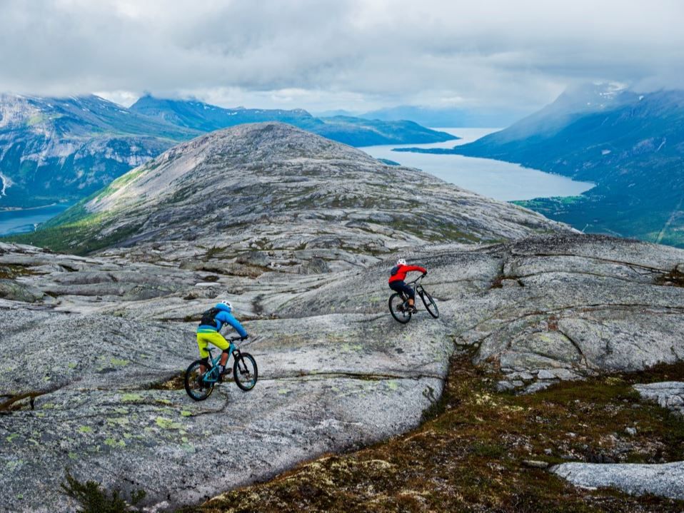 Two mountain bikers cycle over a rocky mountain plateau with fjords and blue mountains under cloudy skies.