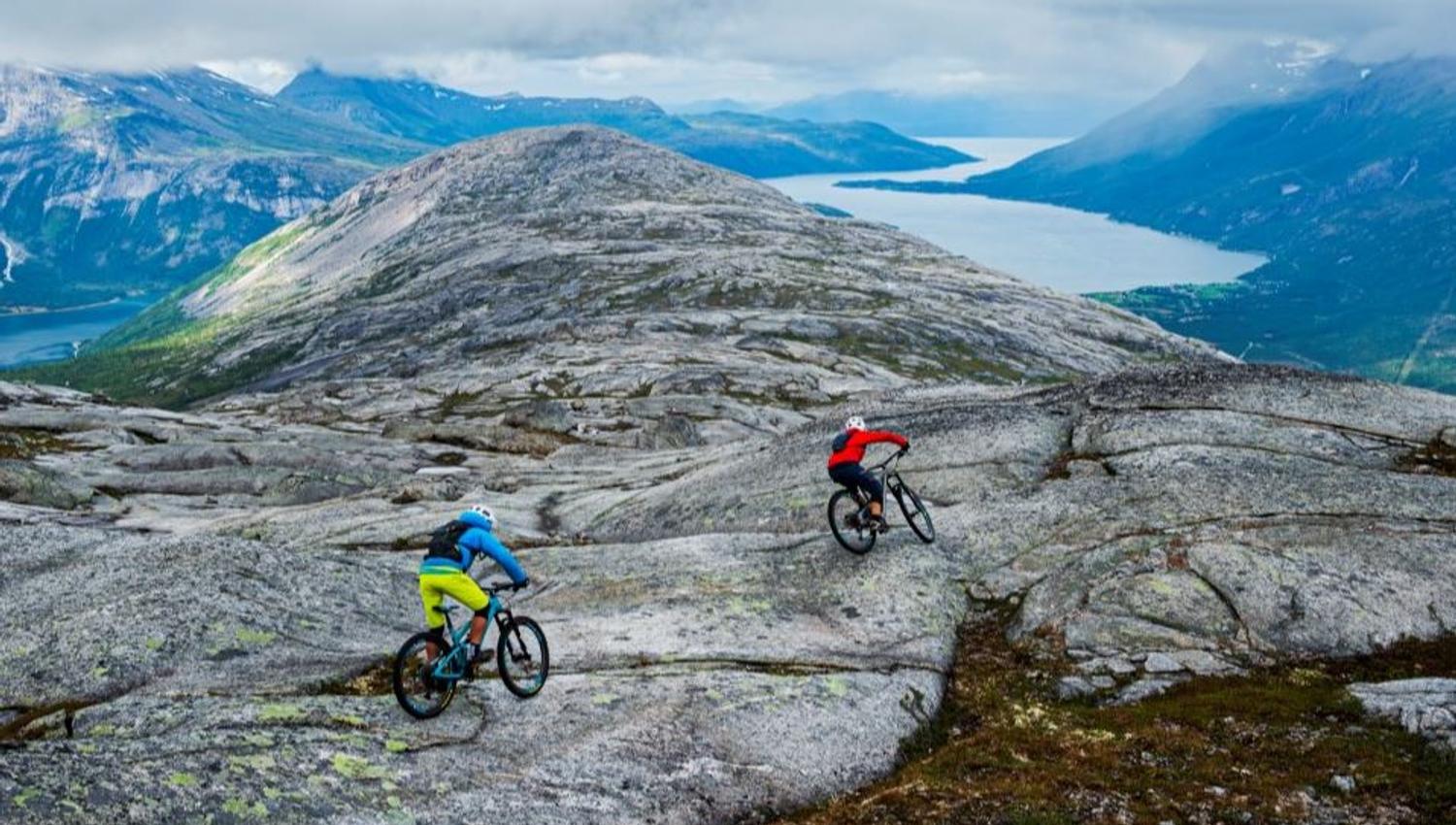 Two mountain bikers cycle over a rocky mountain plateau with fjords and blue mountains under cloudy skies.