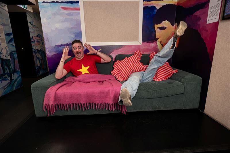 A person laughs while lying on a sofa in a colourful museum room, with legs raised and hands in the air.