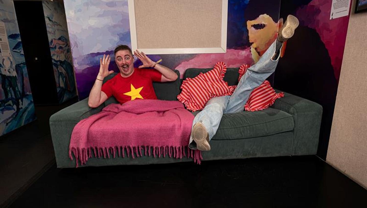 A person laughs while lying on a sofa in a colourful museum room, with legs raised and hands in the air.