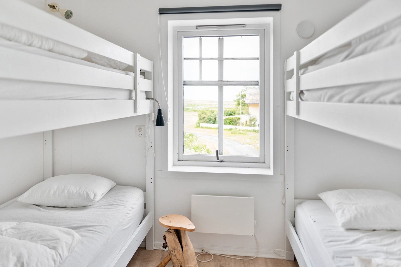 Small white bedroom with two bunk beds, white bedding, a centered window, and a small wooden stool.