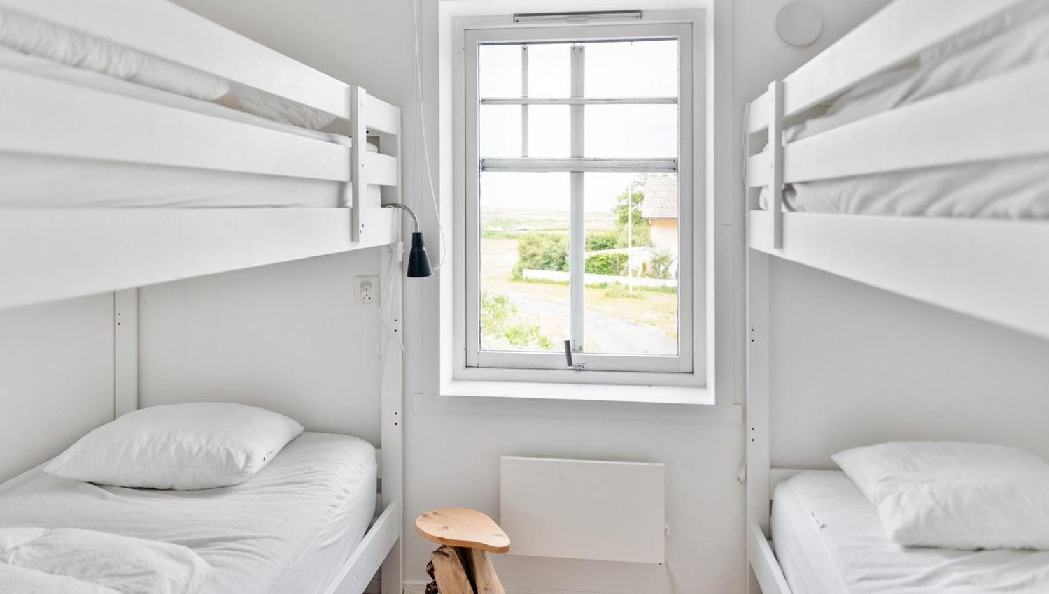 Small white bedroom with two bunk beds, white bedding, a centered window, and a small wooden stool.