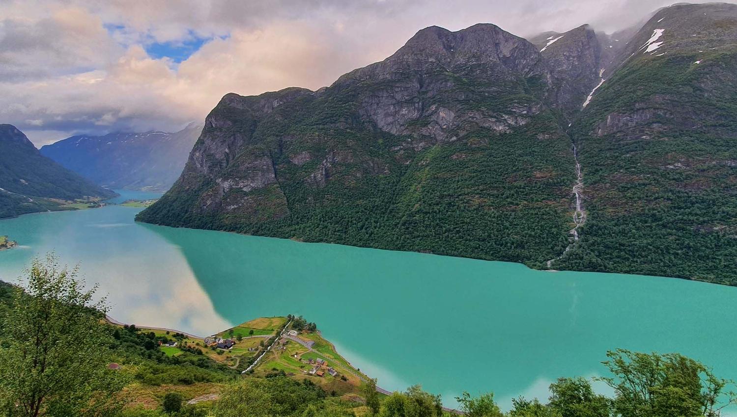 View of a turquoise fjord bordered by steep forested mountains, with a thin waterfall and small farms at shore.
