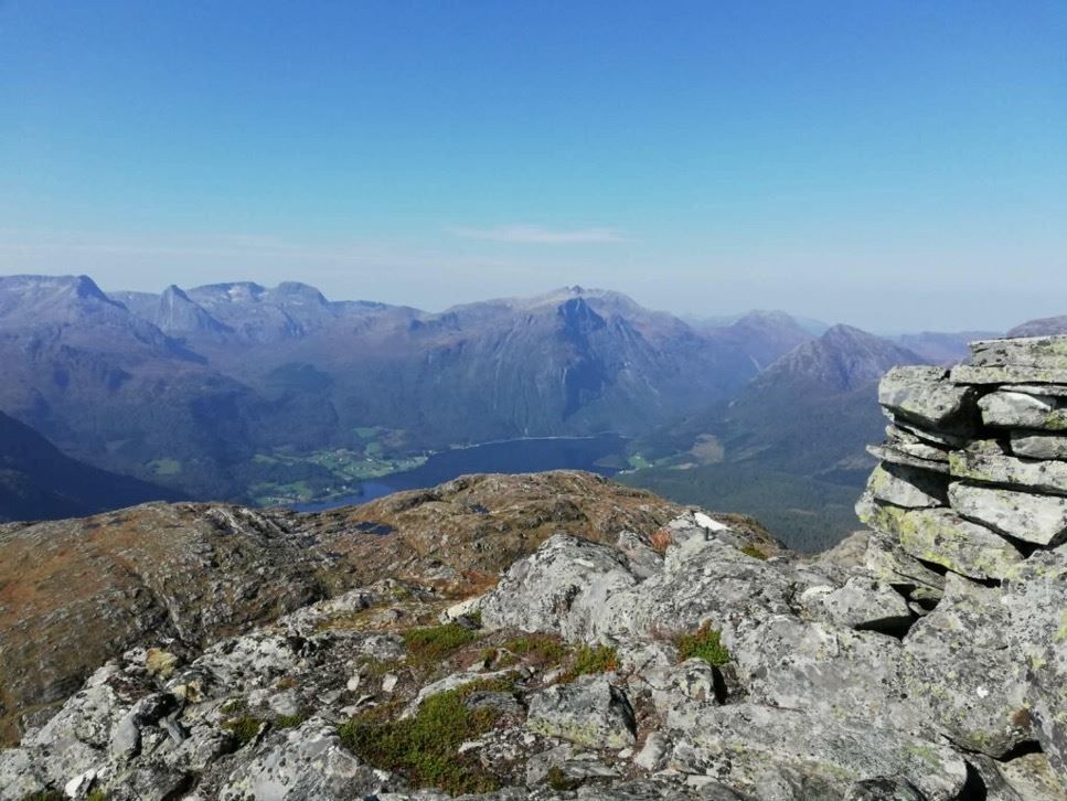 Rocky mountain summit overlooks a valley with a lake, distant rugged peaks, and a clear blue sky..