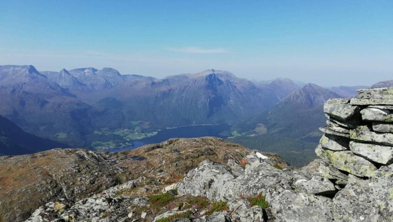 Rocky mountain summit overlooks a valley with a lake, distant rugged peaks, and a clear blue sky..