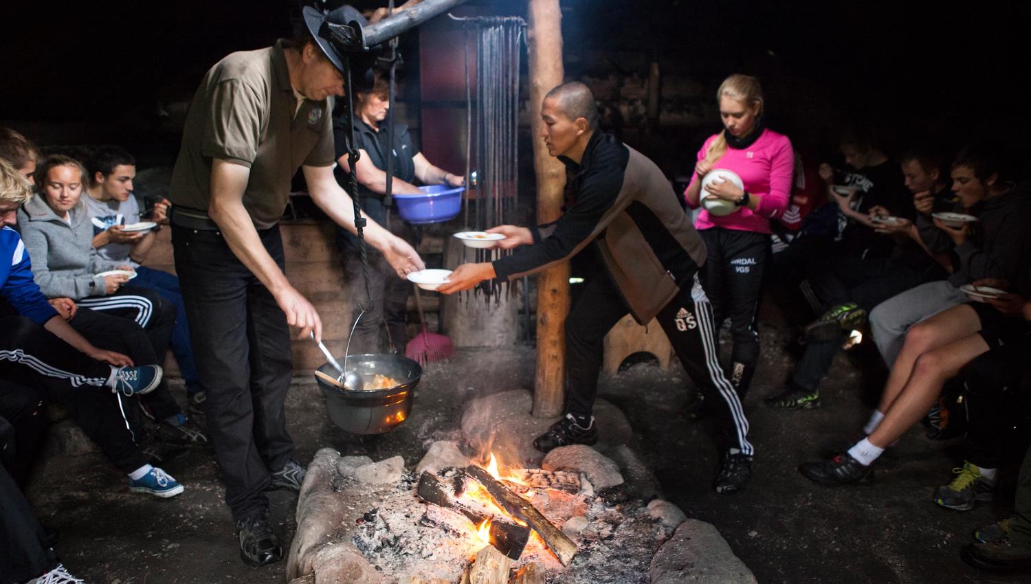Høgetveits Nature Camp. A group of young people is being served food from a pot over a campfire out in the dark. A man wearing a black hat ladles food