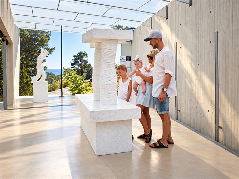 En familie med to barn studerer en hvit marmorskulptur i Midtåsen Skulpturpaviljong, med skog og sjø synlig gjennom glassveggen.