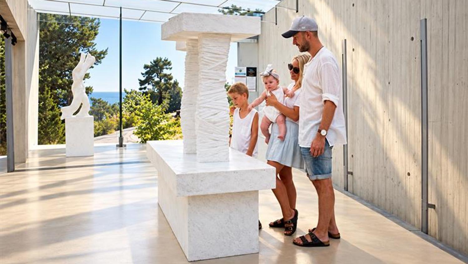 En familie med to barn studerer en hvit marmorskulptur i Midtåsen Skulpturpaviljong, med skog og sjø synlig gjennom glassveggen.