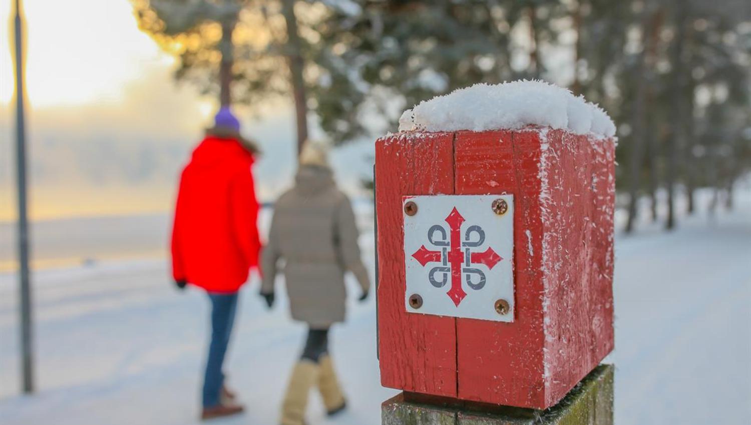 En rød merkestolpe med et hvitt symbol står i snøen, med to personer som går i bakgrunnen.