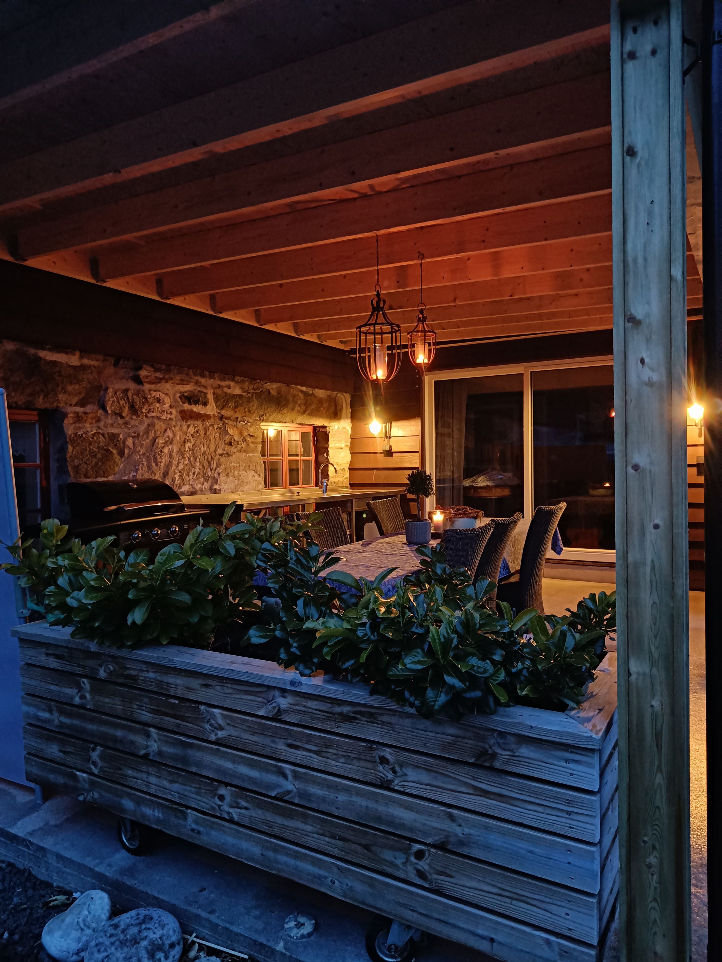 Orheimstunet in Nordfjord - Covered outdoor patio with wooden beams, hanging lanterns, dining set, grill, and planter, lit at dusk..