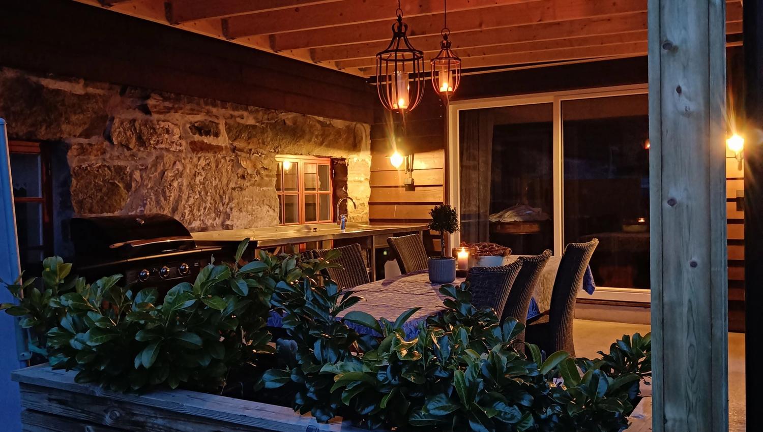 Orheimstunet in Nordfjord - Covered outdoor patio with wooden beams, hanging lanterns, dining set, grill, and planter, lit at dusk..