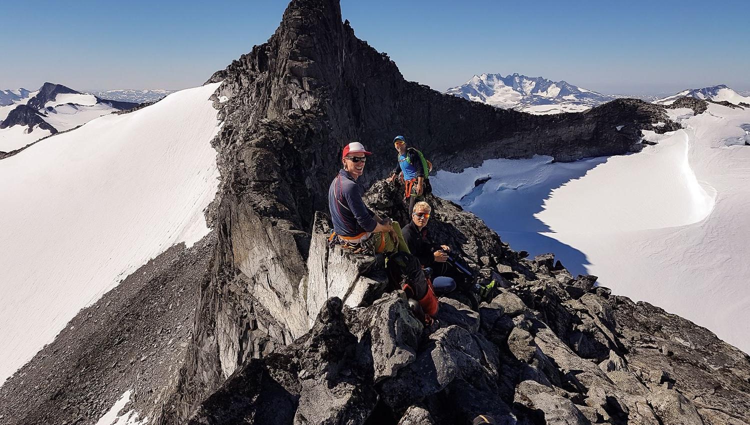Drie klimmers in helmen en harnassen zitten op een rotsachtige alpenkam boven sneeuwvelden onder een heldere blauwe lucht..