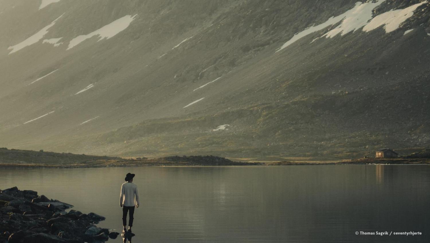 Ein person står ved steinstrand og ser utover eit roleg fjellvatn med bratte fjellsider og snøflekker i bakgrunnen..