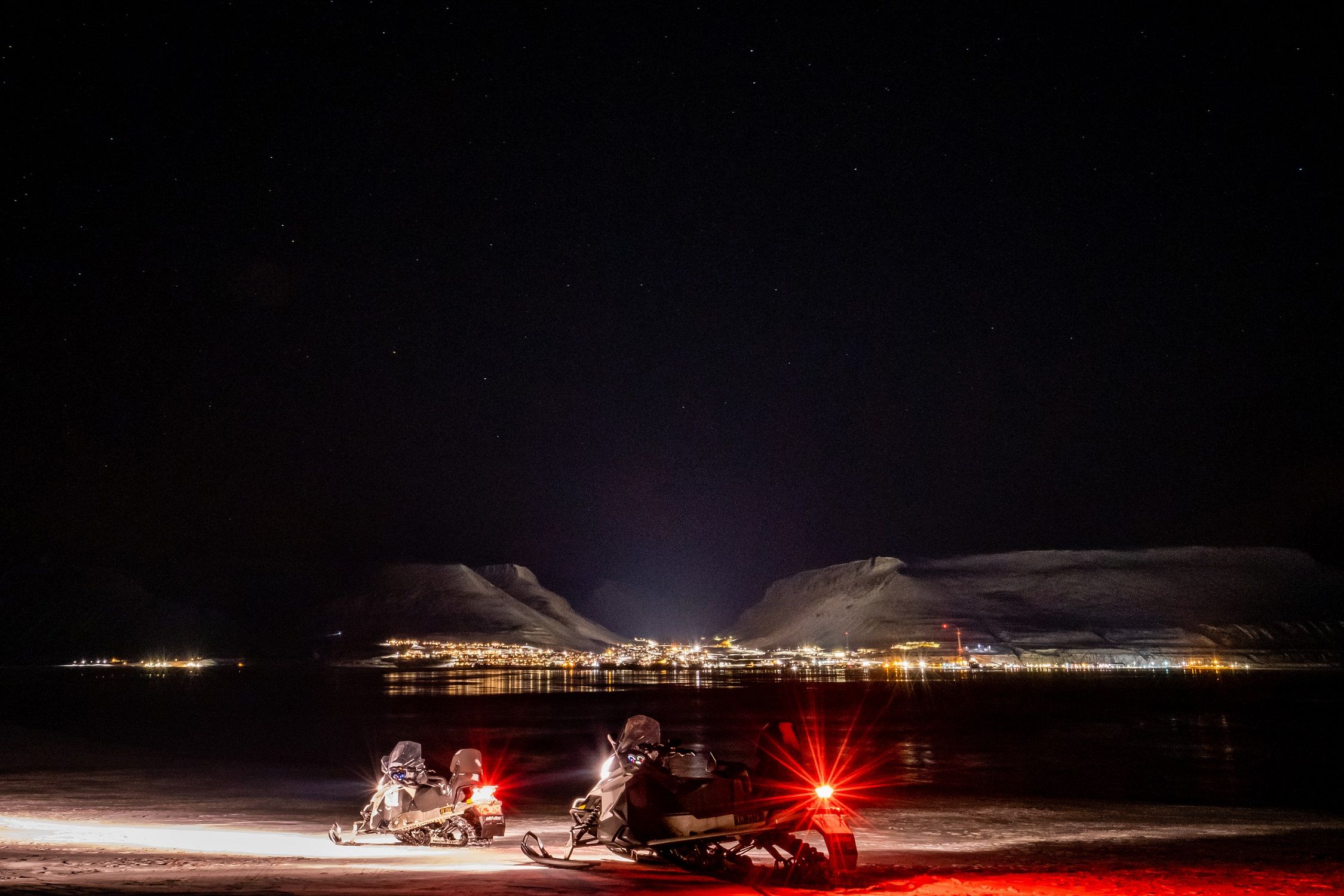 Two snowmobiles in the foreground with town lights from Longyearbyen in the background surrounded by the darkness of night
