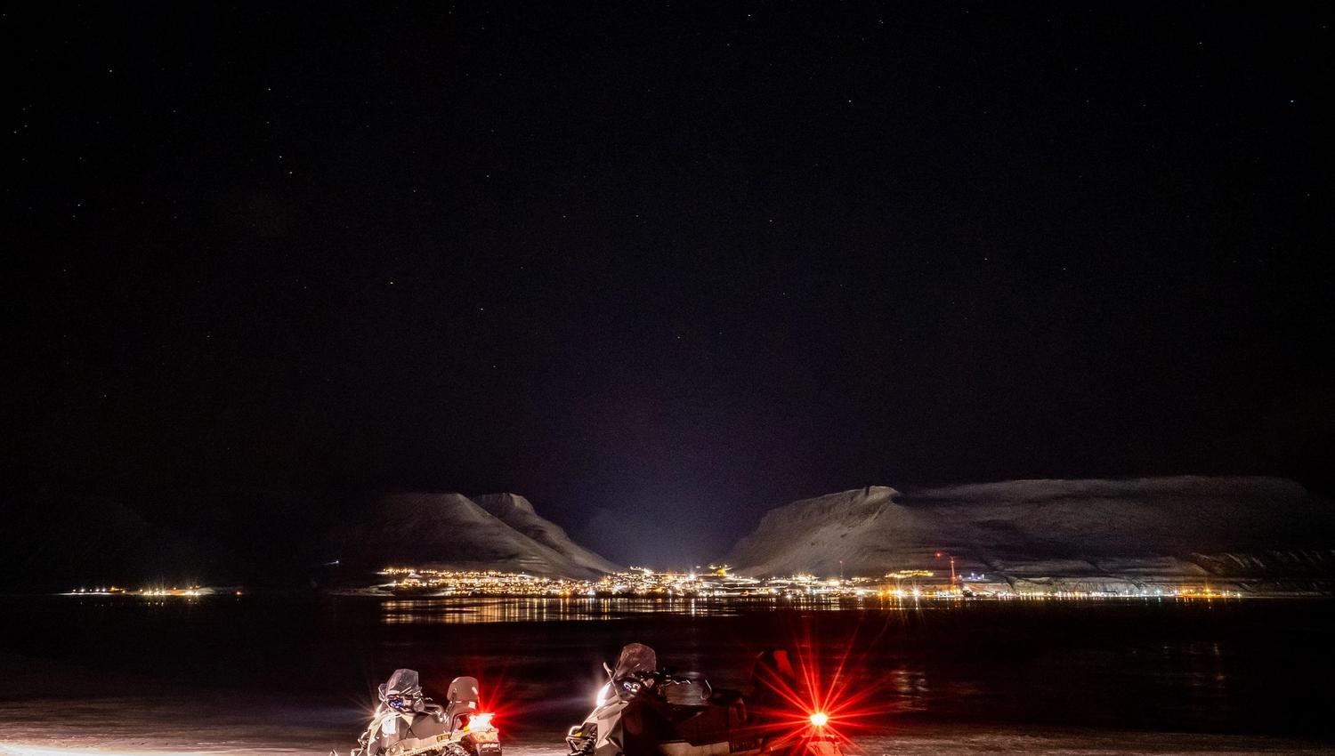 Two snowmobiles in the foreground with town lights from Longyearbyen in the background surrounded by the darkness of night