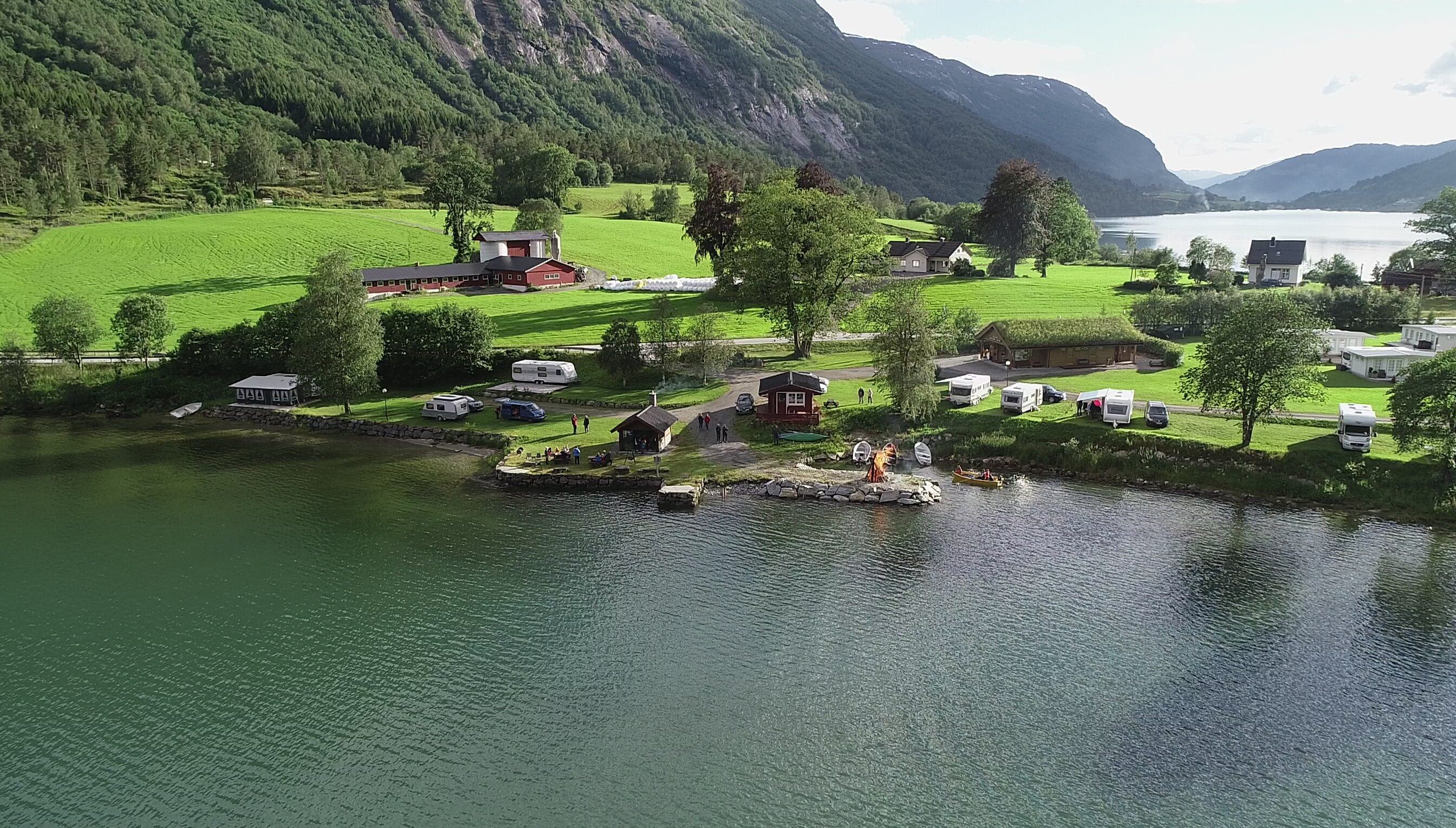 Aerial view of Mindresunde Camping with campers, small cabins, parked RVs, boats, and green fields below forested mountains in Stryn.