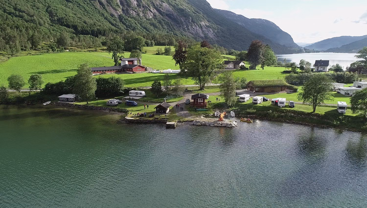 Aerial view of Mindresunde Camping with campers, small cabins, parked RVs, boats, and green fields below forested mountains in Stryn.