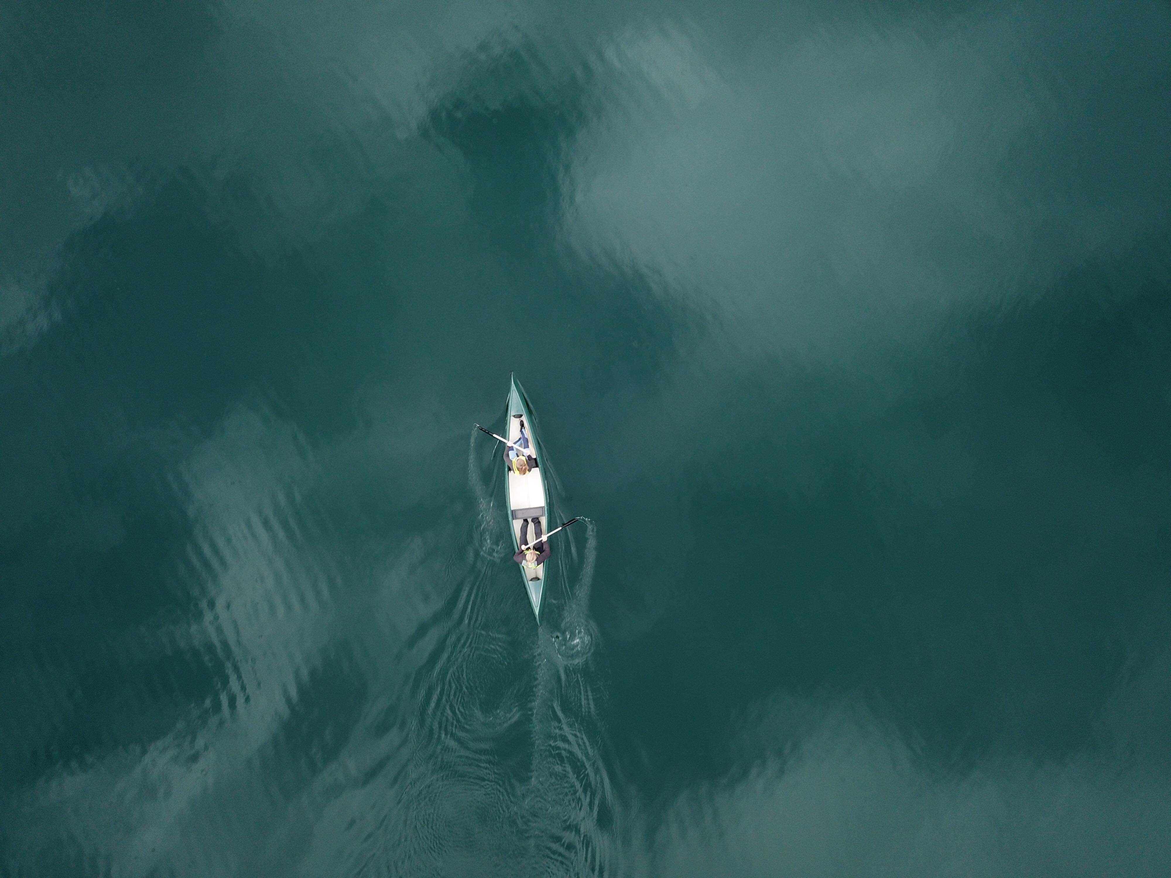 Aerial view of a narrow rowing boat with two people using oars, leaving ripples on dark green water.