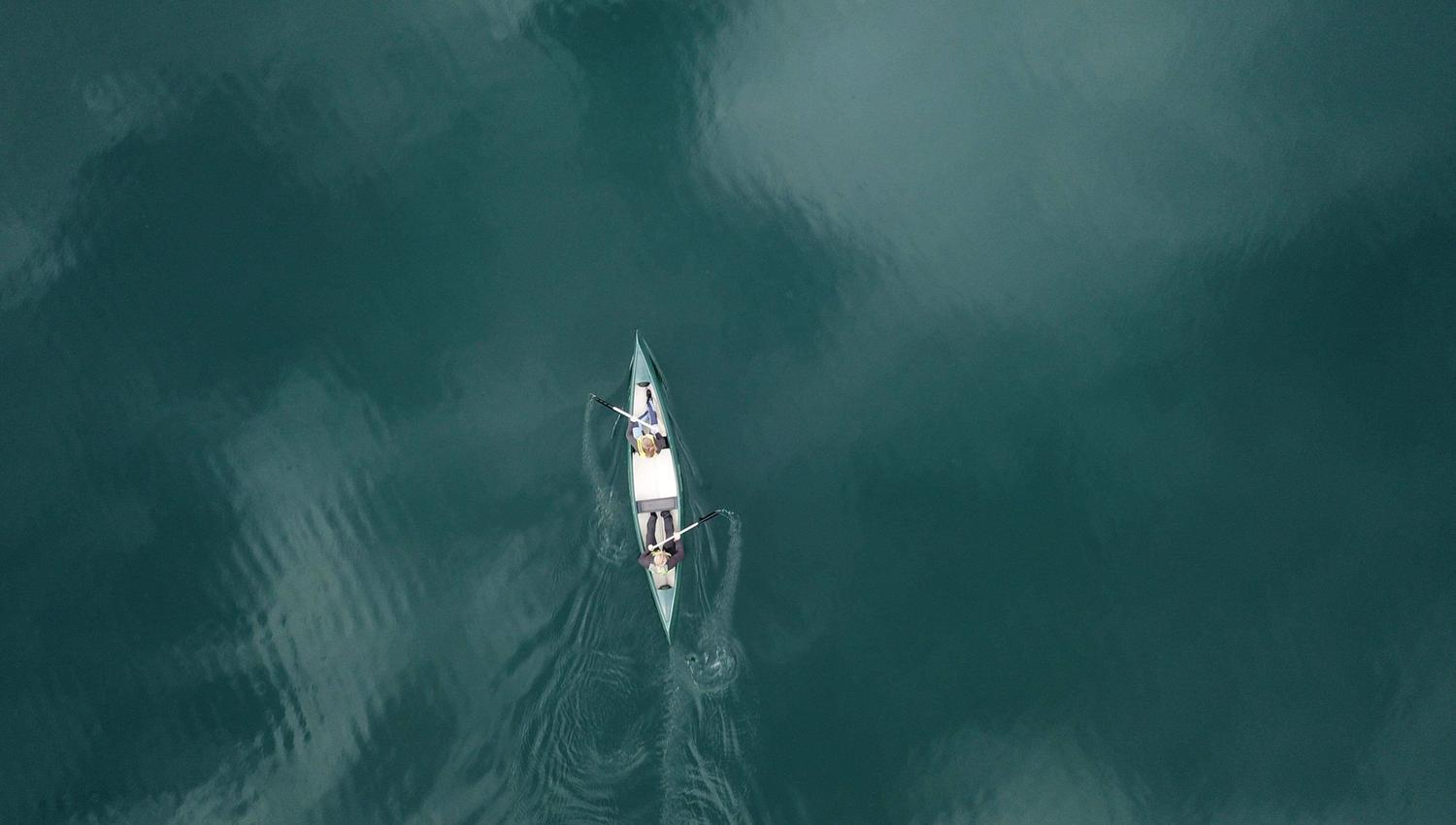 Aerial view of a narrow rowing boat with two people using oars, leaving ripples on dark green water.