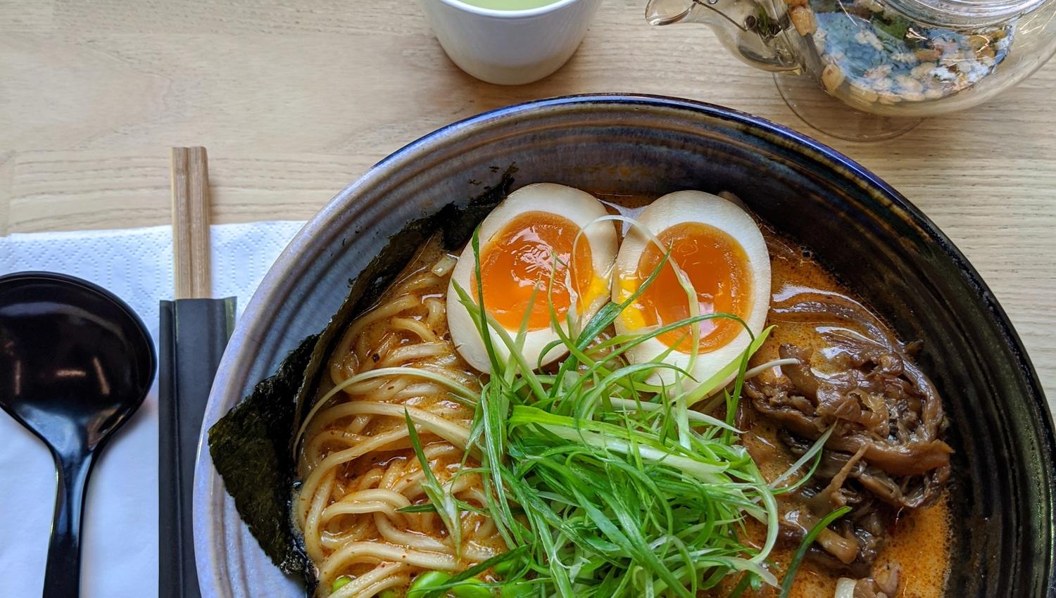 Detaljbilde av ramen i en sort skål, ved siden av bestikk og en kopp grønn te.