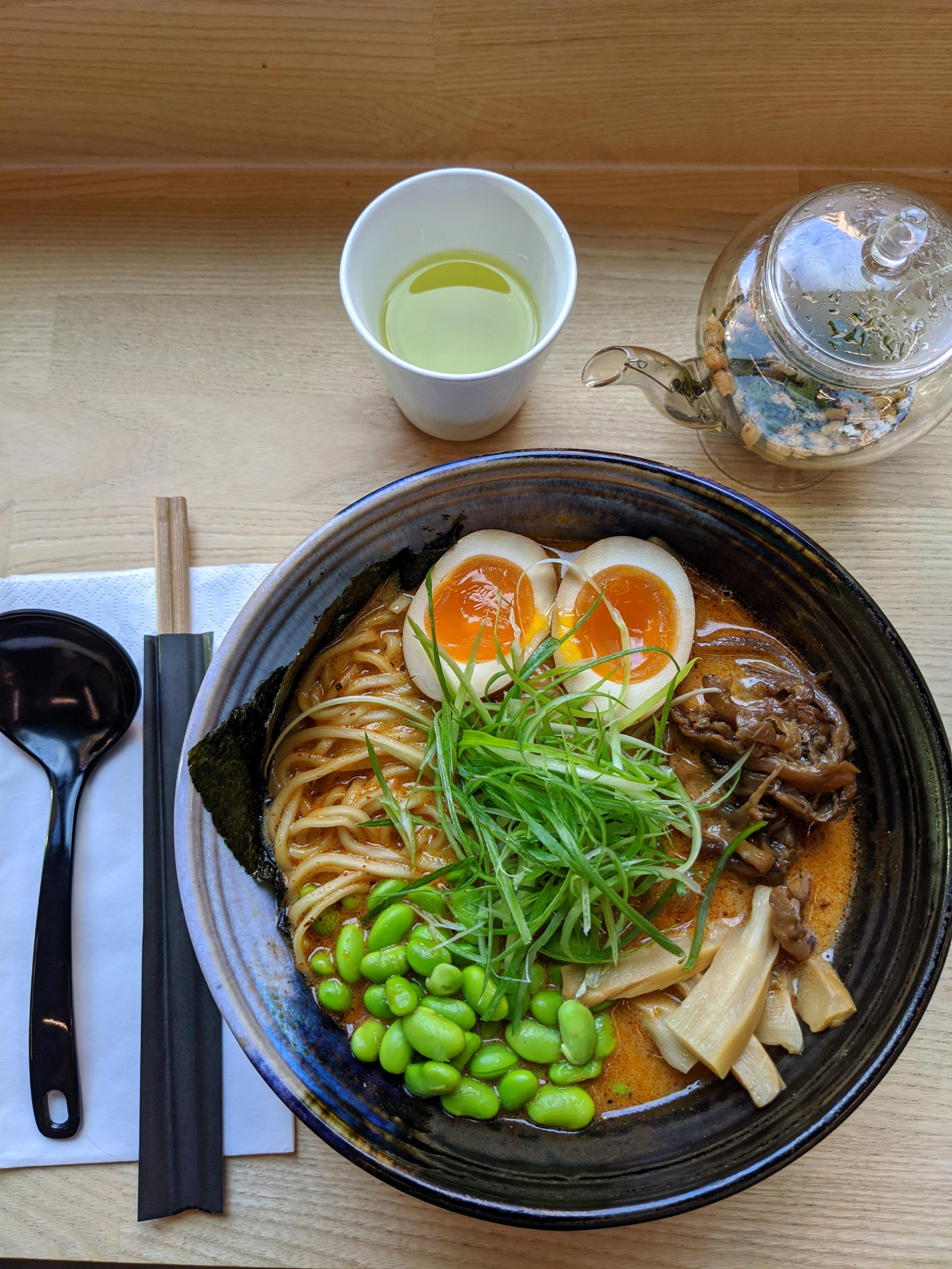 Detail photo of ramen in a black bowl, next to some cutlery and a cup of green tea.