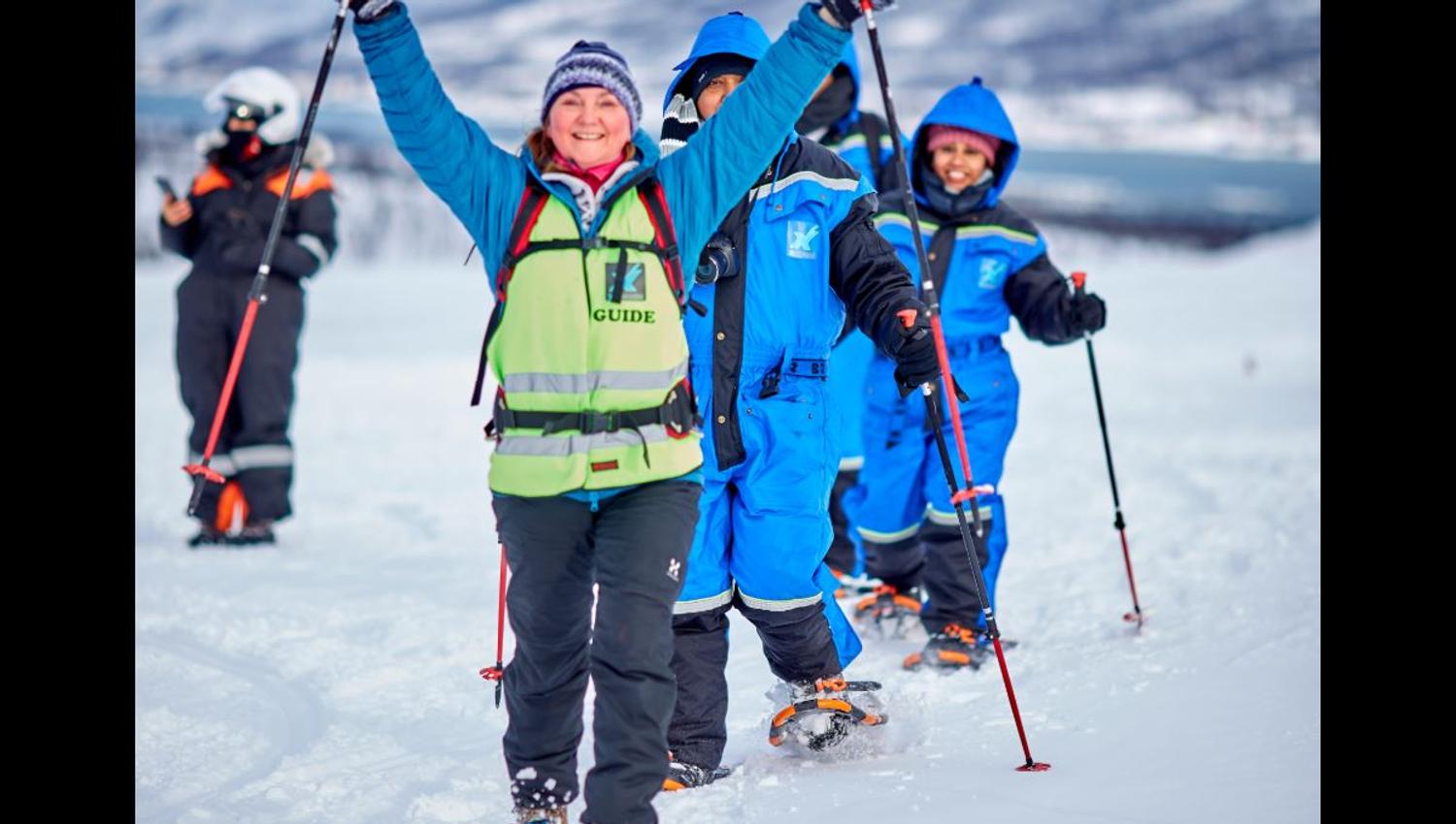 Four persons walking on snowshoes. In the background you can see a person with a thermal suit and a helmet.