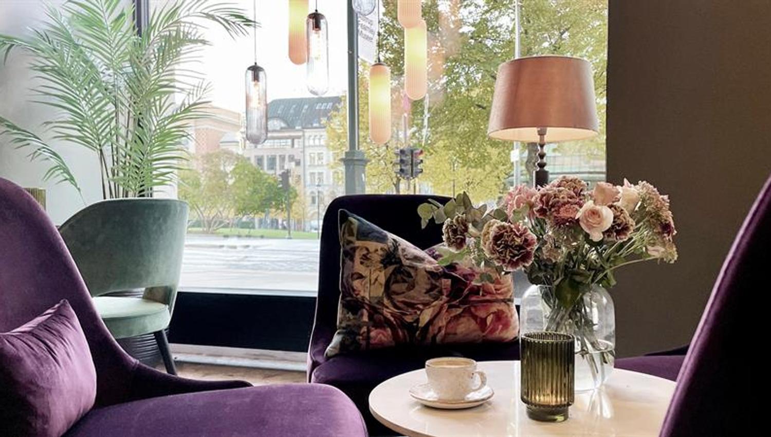 Resepsjonsområde med lilla lenestoler, lite bord med blomster i en potte, telys og kaffekopp, og panoramavindu mot Karl Johans gate