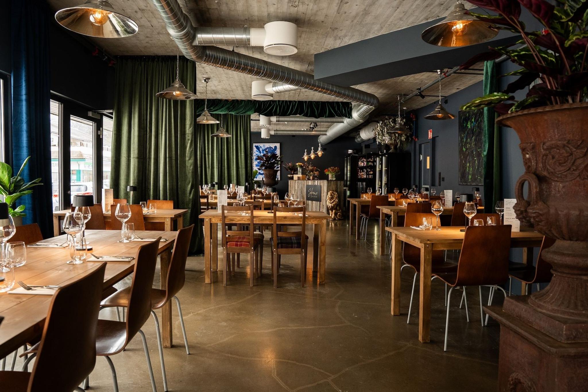 Wide view of a modern restaurant, Sølvane Wine & Dine in Gloppen, Vestland, with wooden tables set with glassware, green curtains, and pendant lights..
