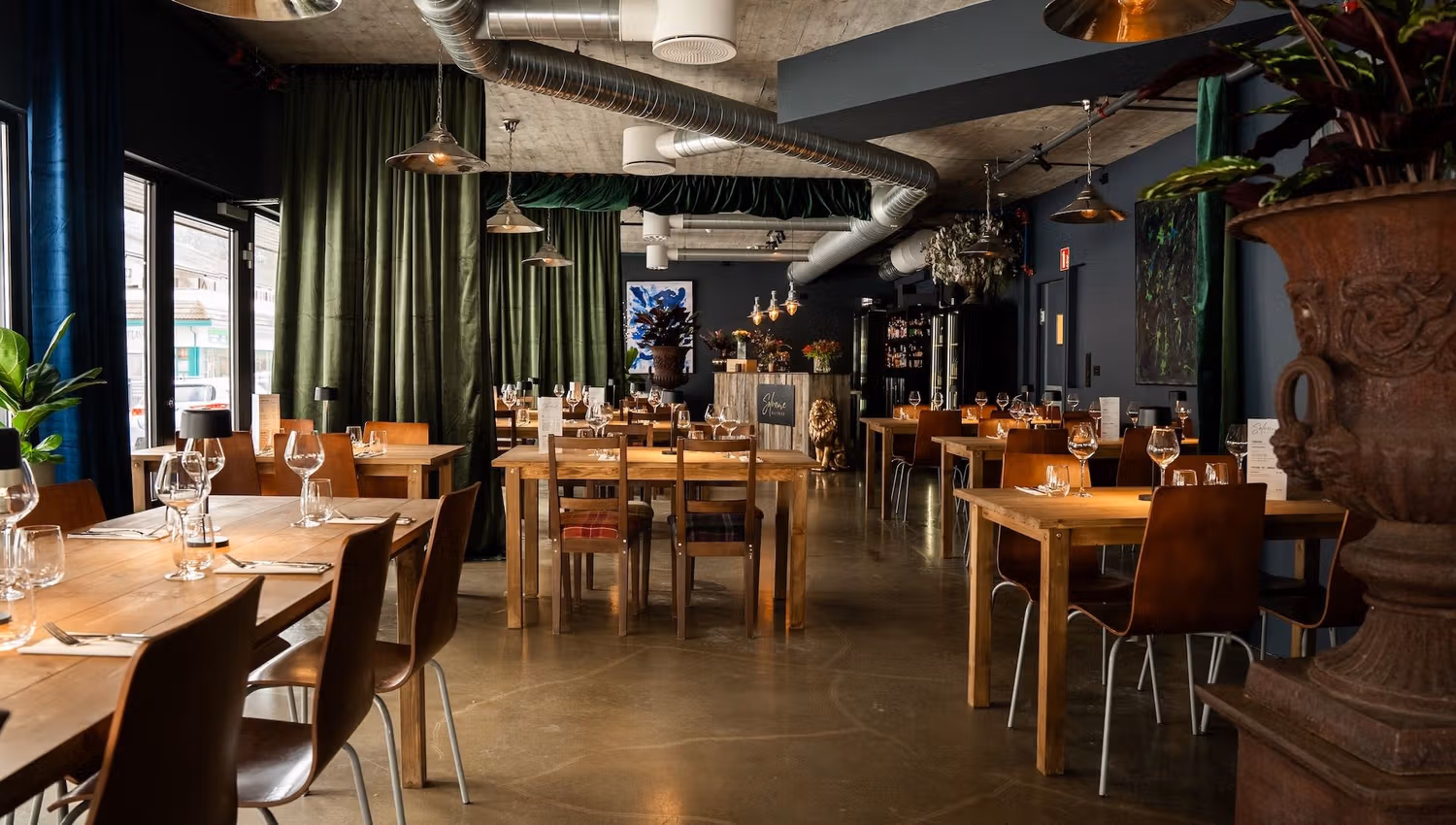 Wide view of a modern restaurant, Sølvane Wine & Dine in Gloppen, Vestland, with wooden tables set with glassware, green curtains, and pendant lights..
