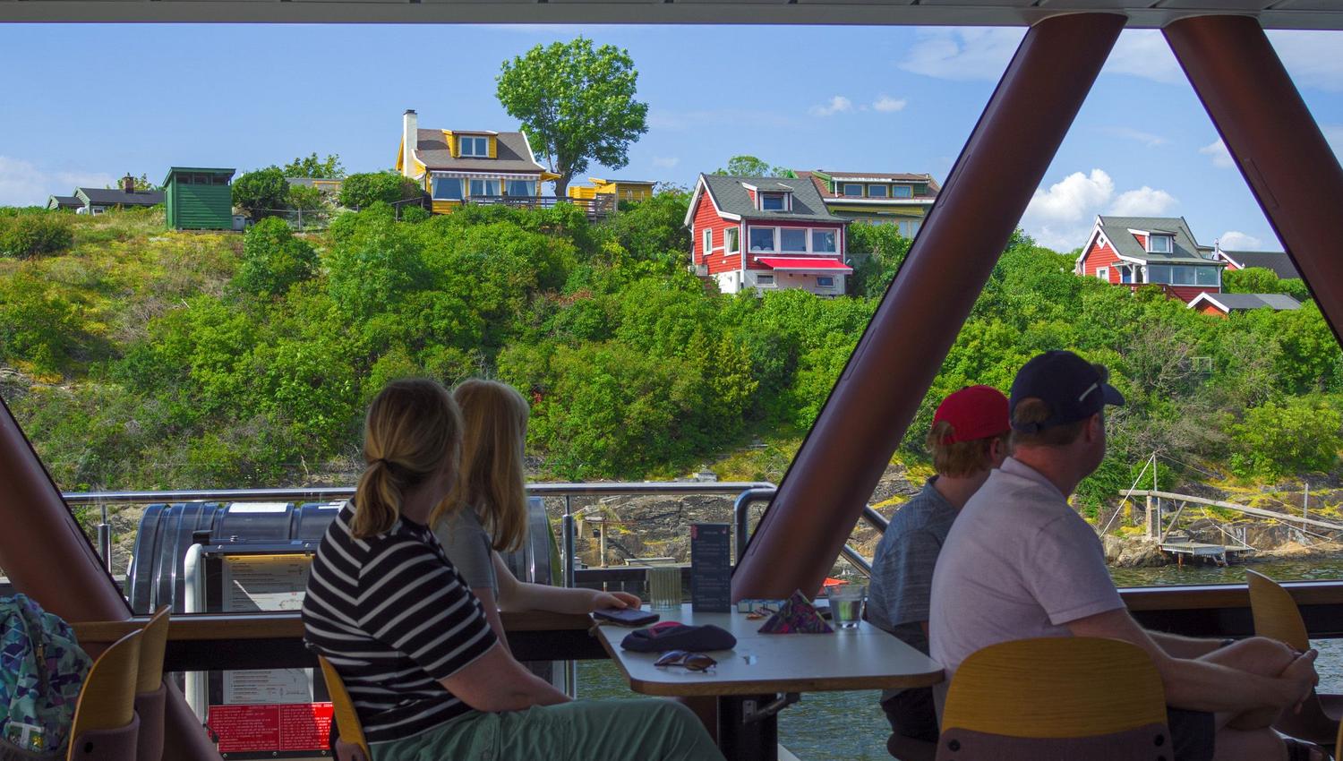 Personer sitter inne i en båt og ser ut gjennom panoramavinduet mot fargerike hus og grønn natur på land.