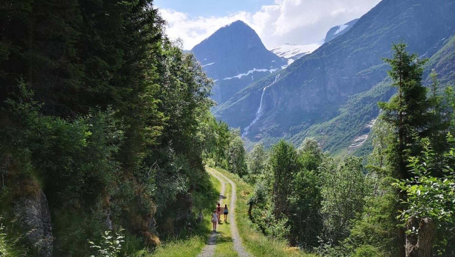 Two hikers walk on a gravel path through green forest, with high mountains and a distant waterfall under clouds in Oldedalen..