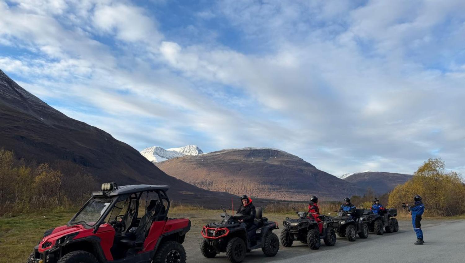 Mange firhjulingen på rekke og rad etter en grusvei. I bakgrunnen kan du se et jorde og høye fjell.