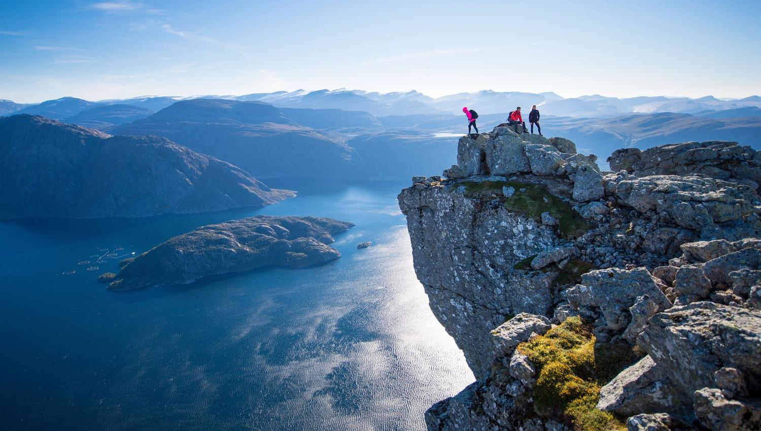 Tre vandrarar står på ein bratt klippe over ein blå fjord, med øyar og fjerne fjell under klar himmel..