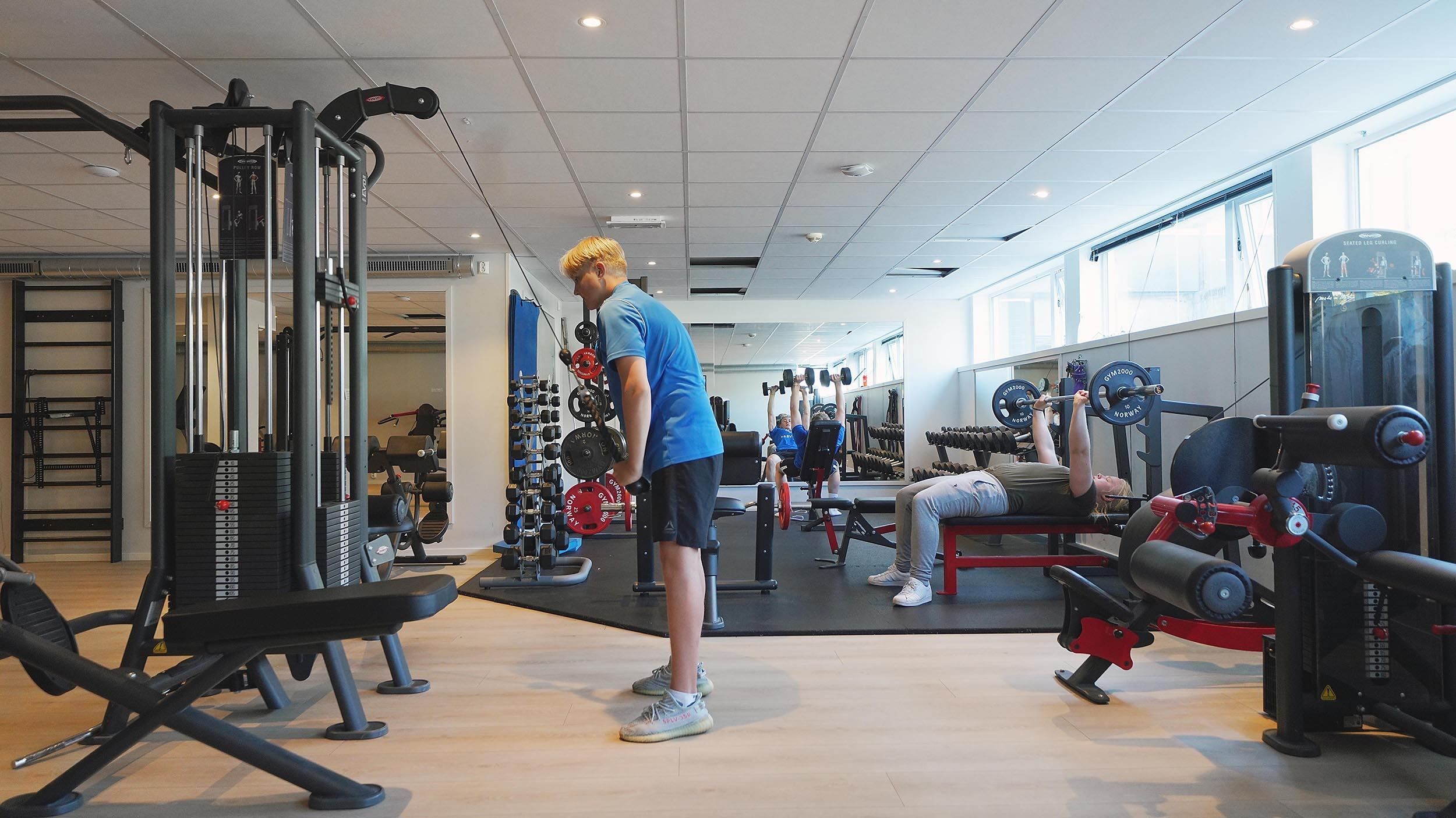 Lyst treningsstudio med apparat og frivekter; ein person står ved kabelmaskin, ein annan benkpressar ved vindauga..