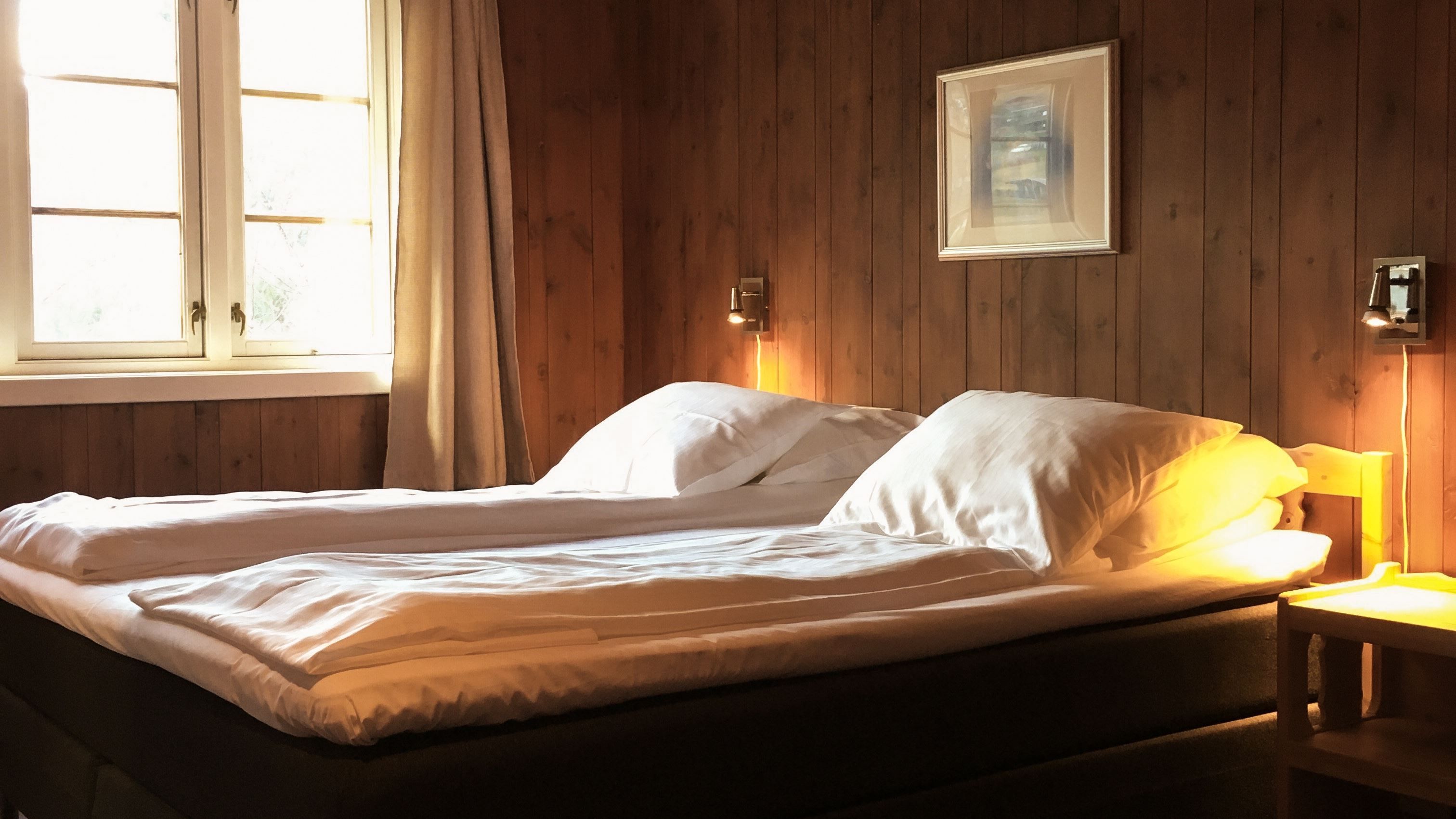 Een met houten lambrisering beklede slaapkamer met een netjes opgemaakt bed, witte linnengoed, warme nachtlampjes en een raam met gordijnen...